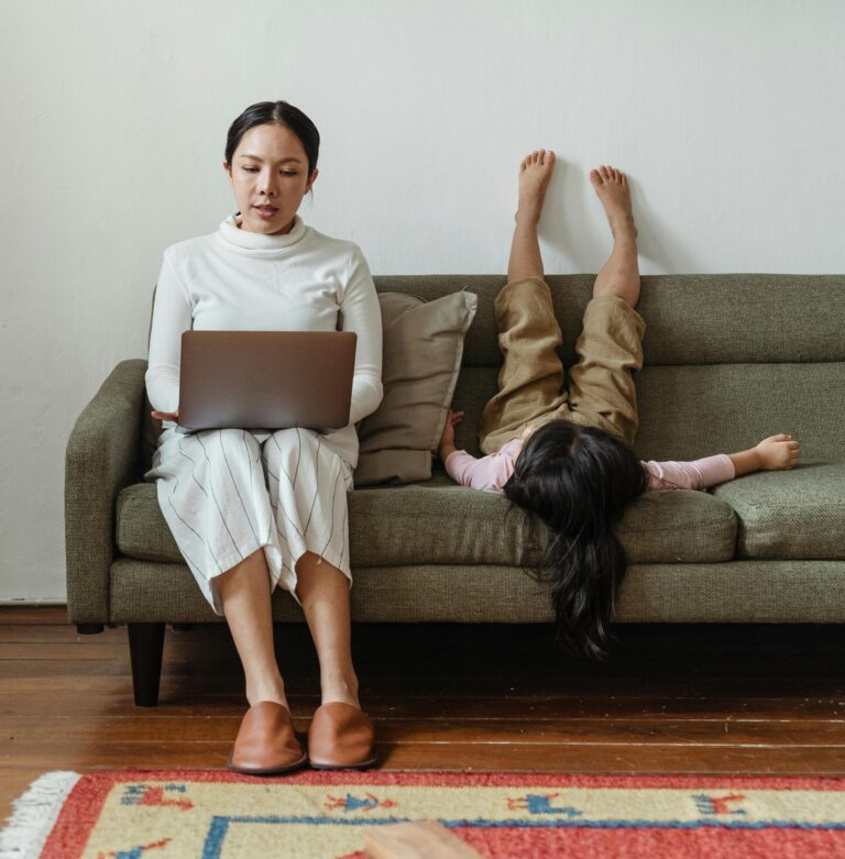 Mother multitasking with laptop while daughter plays on sofa, balancing work and family.